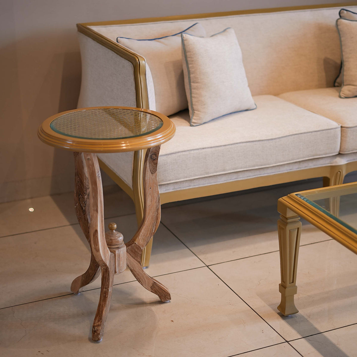 Small round wooden table with glass top next to a beige sofa with cushions on a tiled floor.