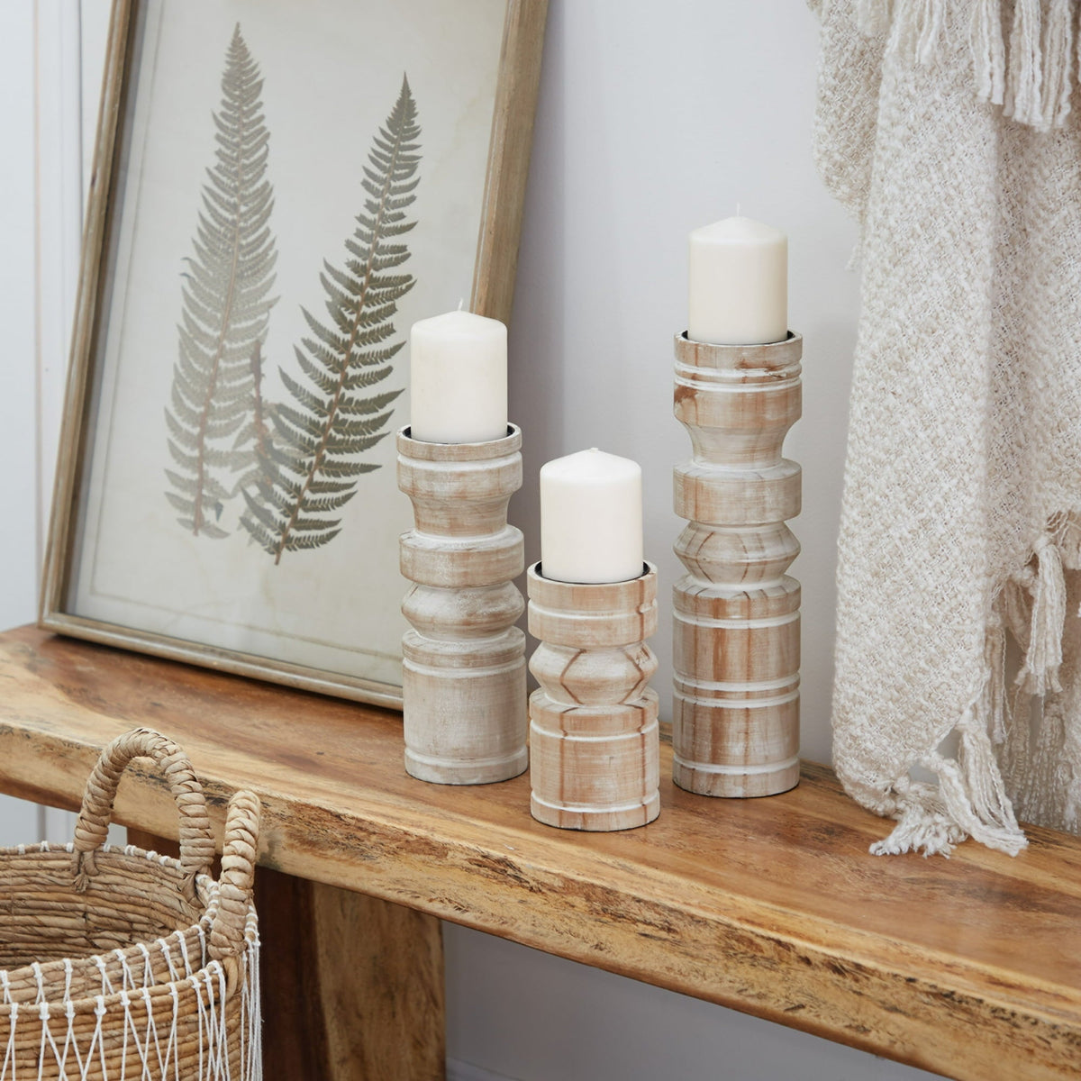 Three white, hand-turned candle stands with a whitewashed patina, displayed on a wooden surface, with a framed picture of a fern and a textured white curtain in the background.