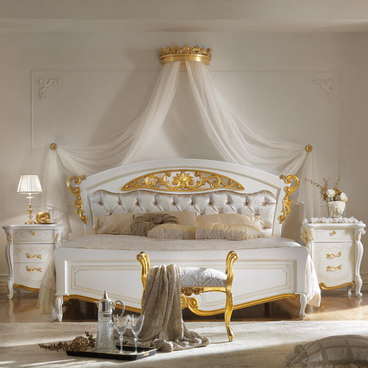 A luxurious white and gold canopy bed with tufted upholstery, draped white fabric canopy, matching nightstands, and a decorative bench in a bright, elegant bedroom.