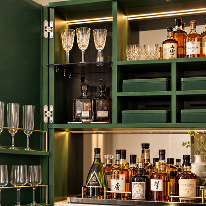 A dark green bar cabinet with open doors, illuminated by warm built-in lighting, filled with wine glasses, bottles, and storage boxes. The cabinet interior and lower shelf are styled with various liquor bottles, crystal glasses, and green storage bins.