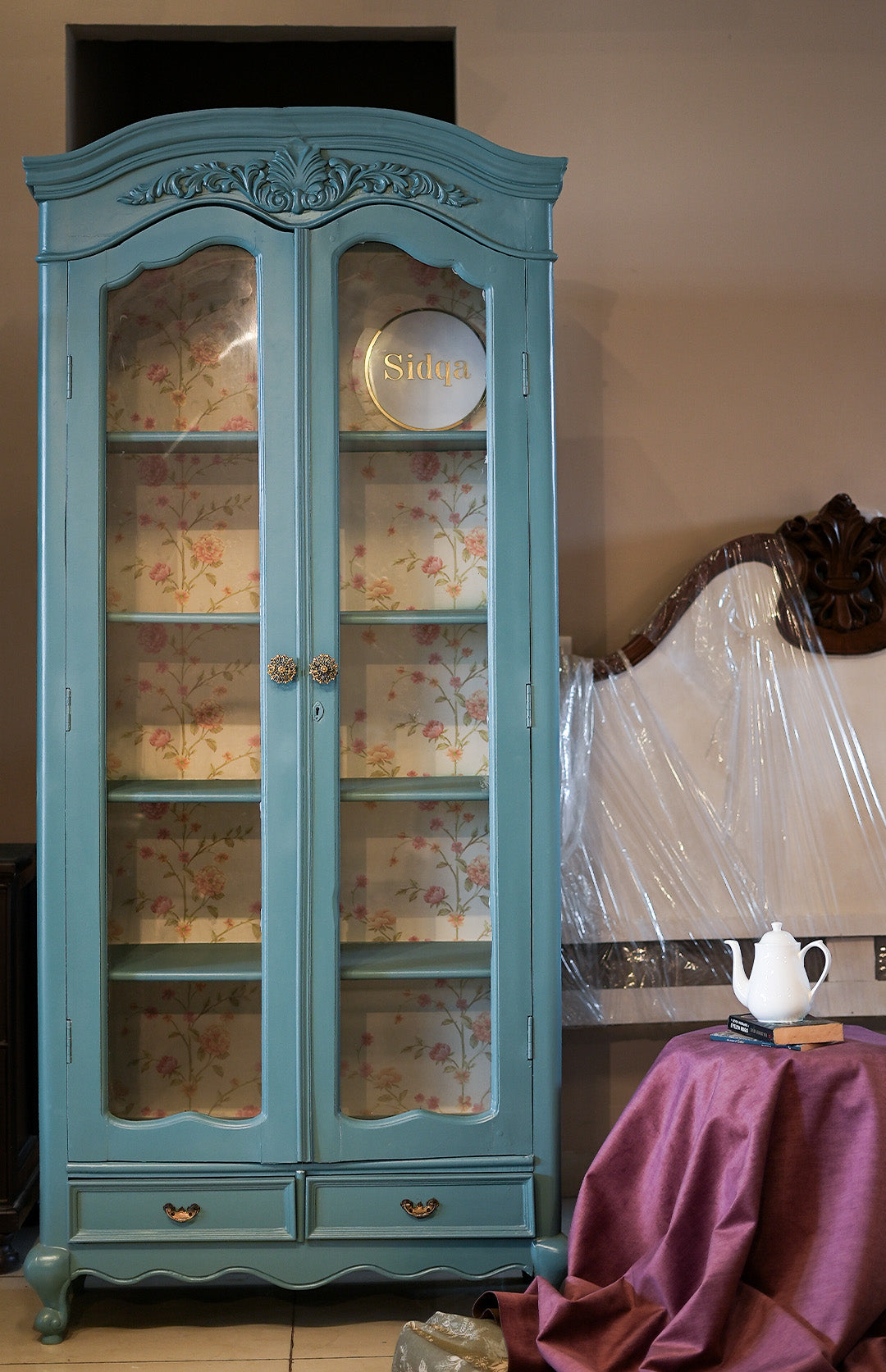 Blue ornate cabinet with floral patterns on glass doors, next to a table with a teapot and books.