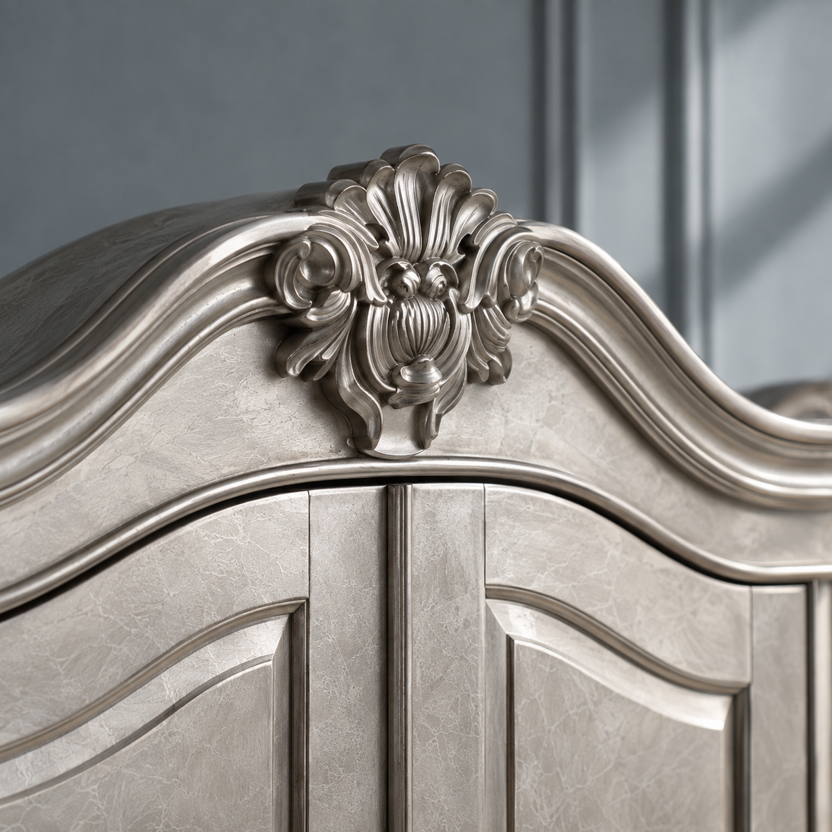 Close-up of an ornate silver bed frame headboard with carved floral details and curved paneling, set against a dark gray paneled background.
