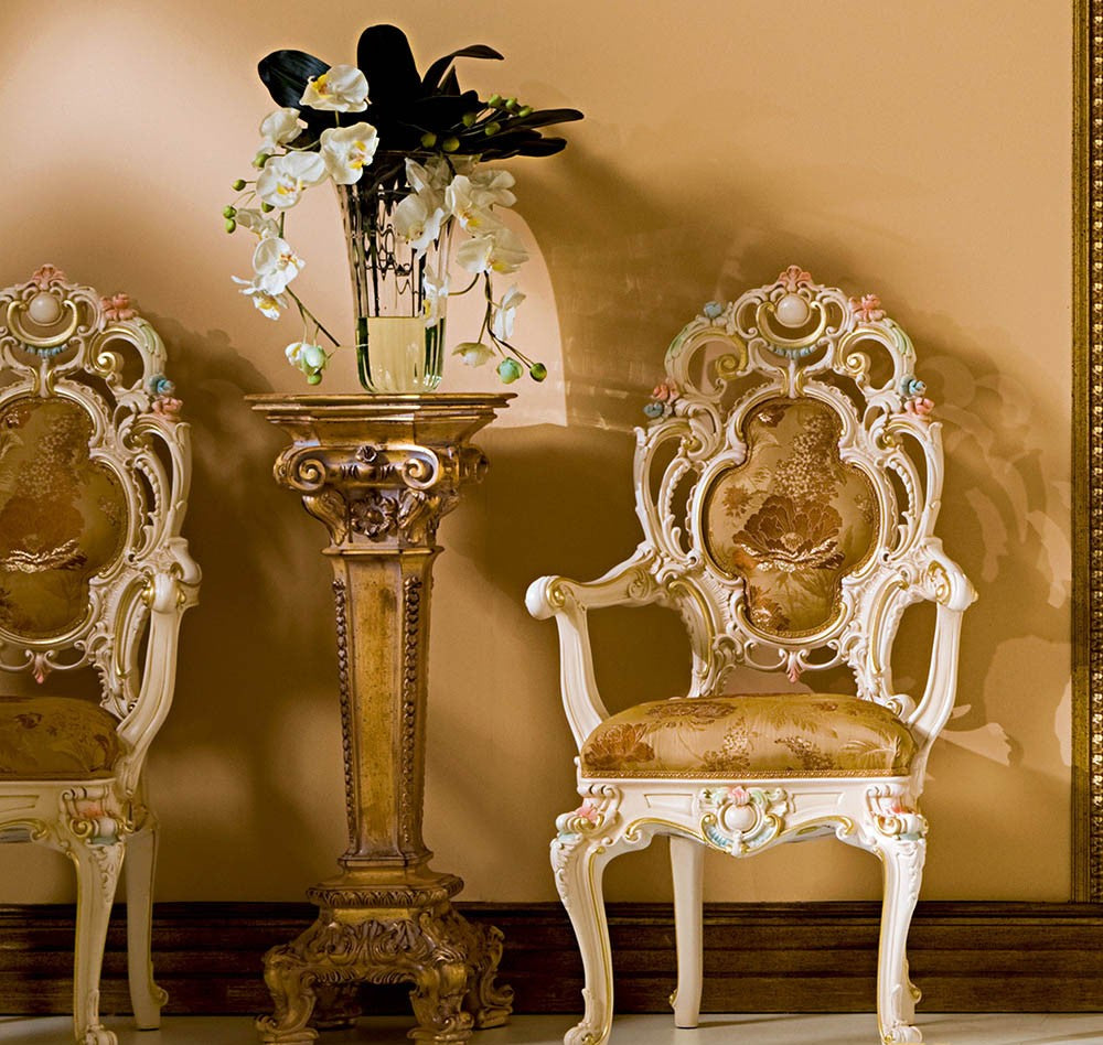 Decorative chairs with intricate designs and a vase of flowers on a pedestal against a beige wall.