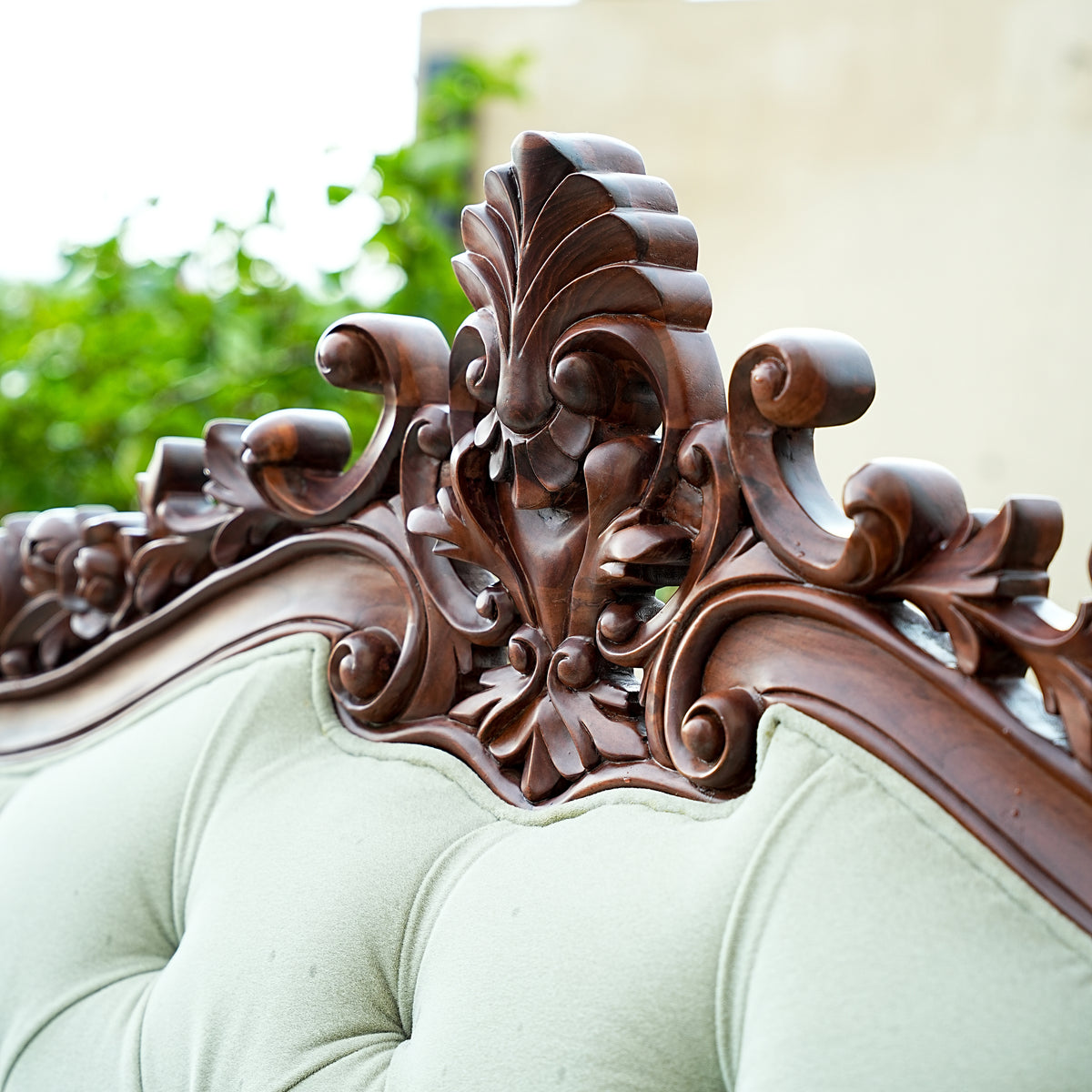 Close-up of ornate wooden carvings on a light green upholstered chair with blurred greenery in the background.