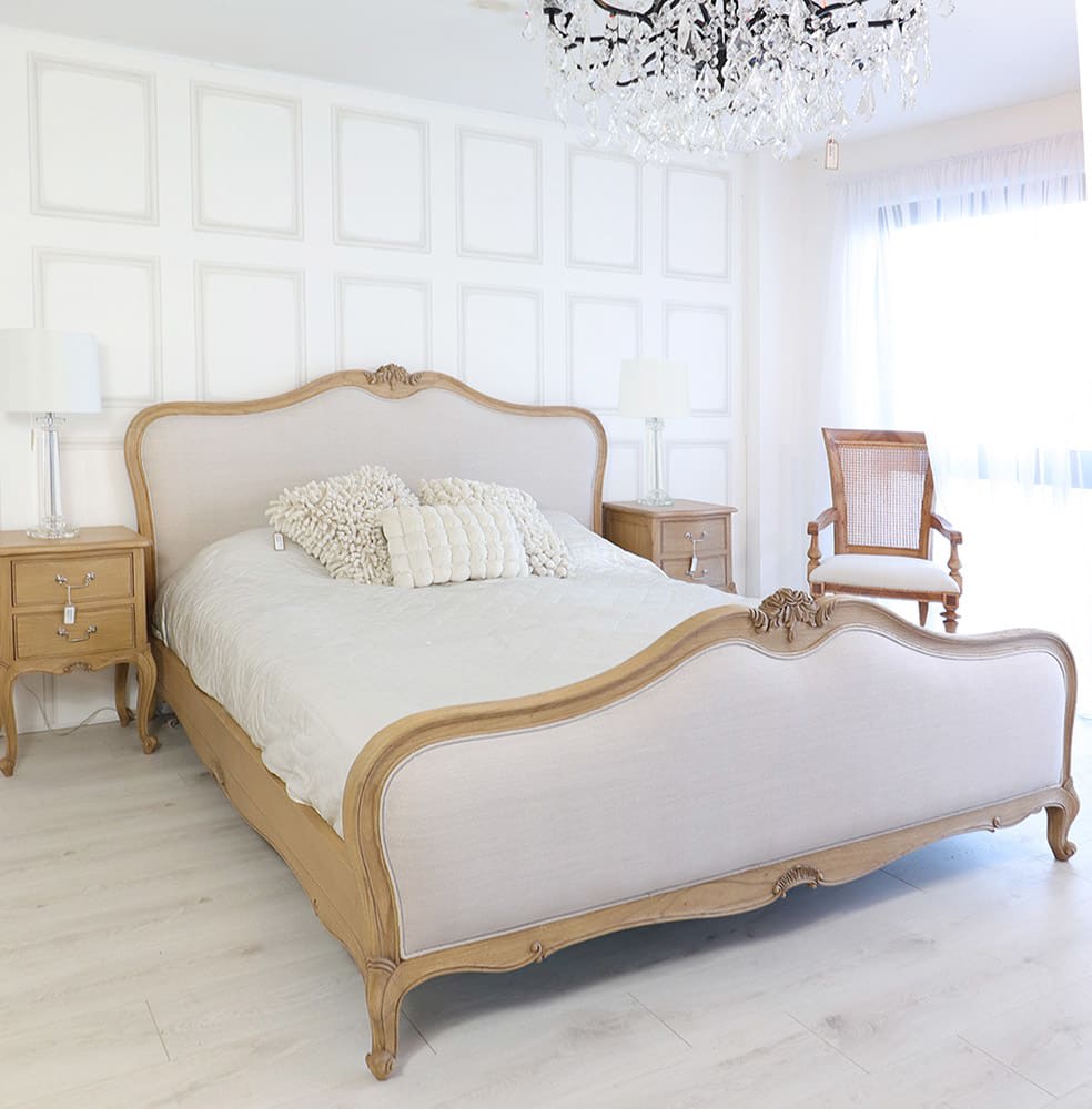 A natural French finish bed with grey upholstery, featuring ornate carvings on the headboard and footboard, placed in a bright room with a chandelier and wooden furniture.