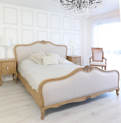 A natural French finish bed with grey upholstery, featuring ornate carvings on the headboard and footboard, placed in a bright room with a chandelier and wooden furniture.