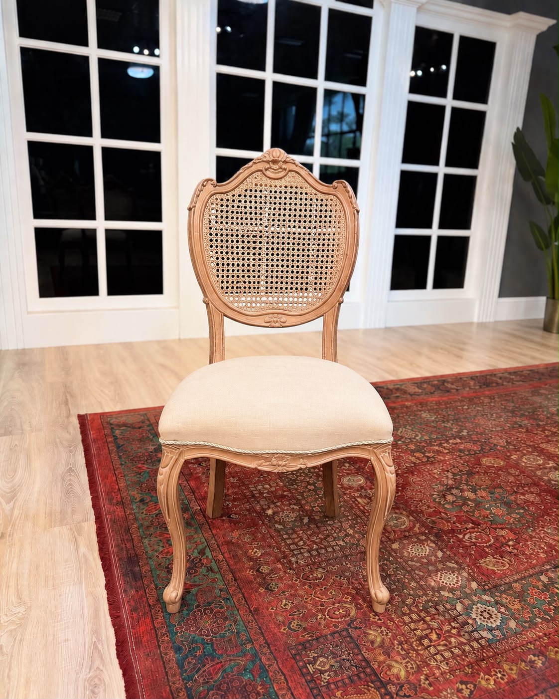A classic French provincial style dining chair with a carved light wood frame, woven cane backrest, and cream upholstered seat cushion, positioned on a red patterned rug in front of white-framed glass doors.