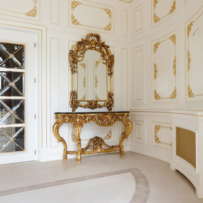 An ornate gold leaf console table with intricate carvings, accompanied by a large mirror, positioned in a room with white walls and decorative accents.