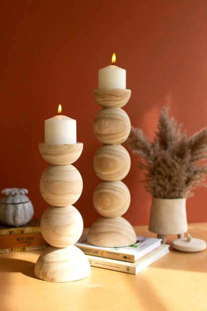 Two cylindrical candle stands made of natural bamboo with lit candles on top, placed on a wooden surface against a red wall. One stand is taller than the other.