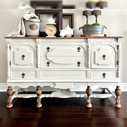 An antique ivory sideboard with natural wood top, featuring scalloped aprons, carved corners, and vintage-style pulls. The turned legs are touched with a worn gold finish.