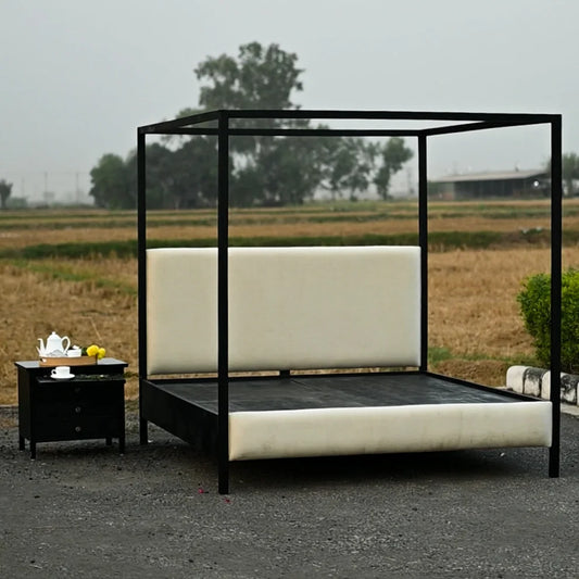 A black metal canopy bed with cream upholstered headboard and footboard, shown outdoors on a gravel surface next to a matching black nightstand with a teapot and lemons. The background is a blurred field and trees under an overcast sky.