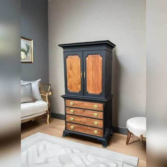 A statueque silhouette of a tallboy armoire with black framing and natural wood grain, featuring brass handles and a mix of upper cabinet and lower drawers.