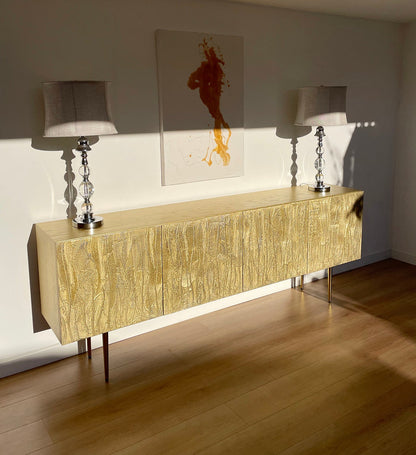 Gold sideboard with two lamps and a piece of abstract art on the wall.