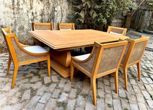 A 6-seater dining set with a wooden table and cane chairs arranged on a cobblestone surface with greenery in the background.