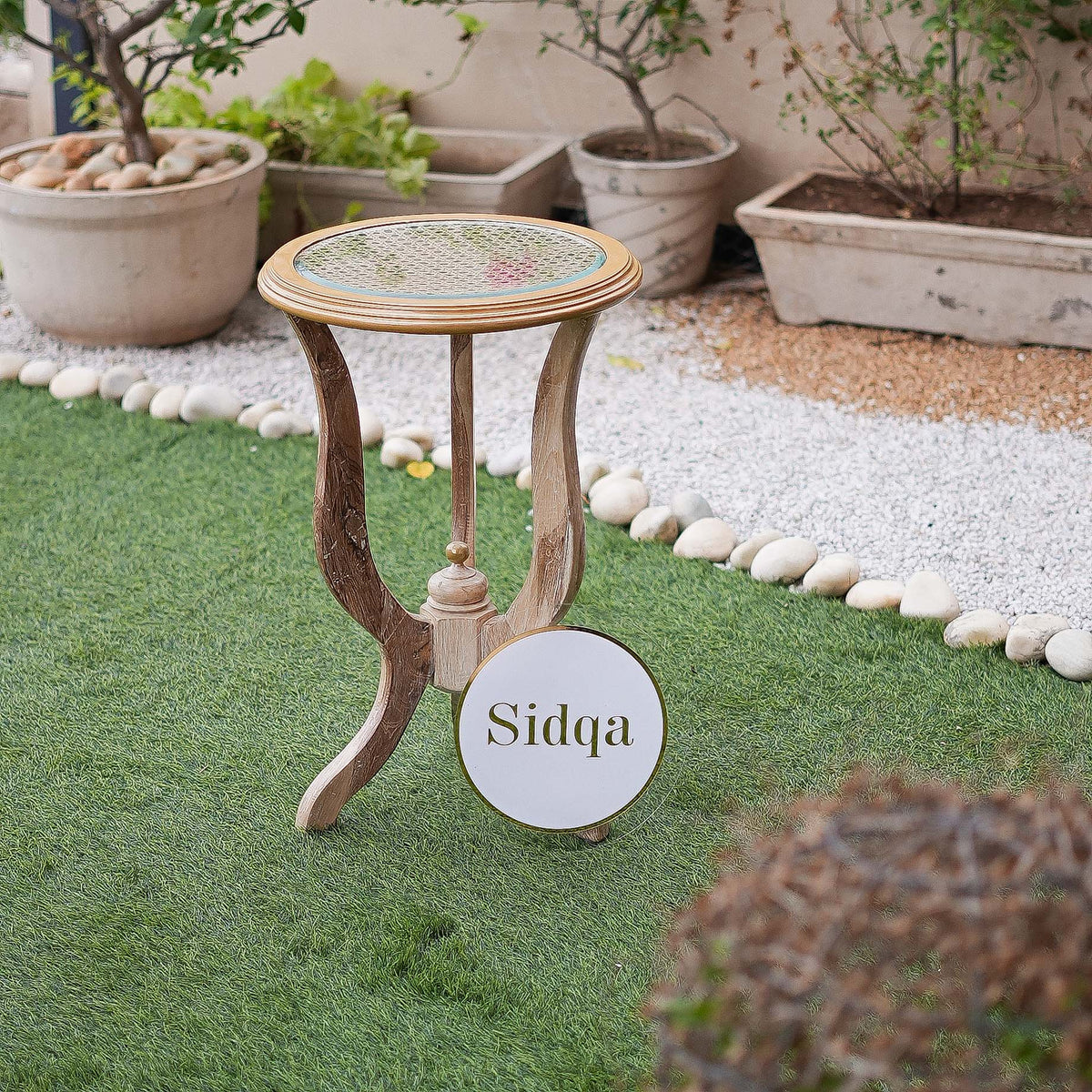 Small round wooden table with decorative top on a grassy area with potted plants in the background