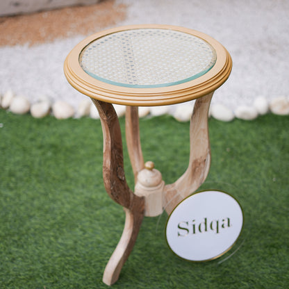 Small round table with wicker top and wooden base on a green surface, branded 'Sidqa'.