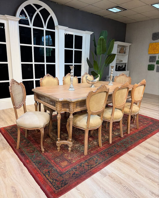 Wooden dining table with chairs on a patterned rug in a room with large windows.