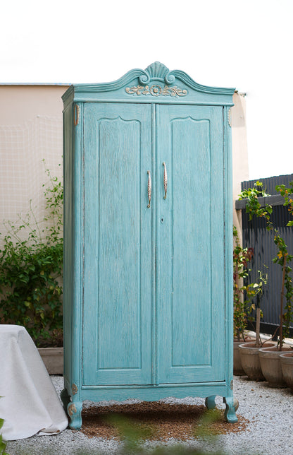 Vintage-style teal wardrobe with decorative top against a white wall.