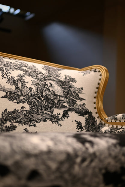 Close-up of a tufted upholstered chair or sofa arm with black toile de Jouy pattern fabric and gold nailhead trim on a dark background.