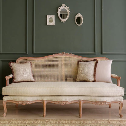 A luxurious beige upholstered 3-seater sofa with cane detailing and teak wood legs, accompanied by two patterned pillows, positioned against a dark green wall with decorative mirrors above.