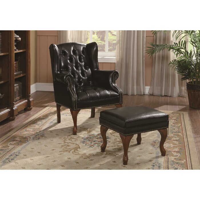 A black upholstered armchair with tufted backrest and ottoman, placed on a patterned rug with mahogany wooden legs in a room with bookshelves and curtains.