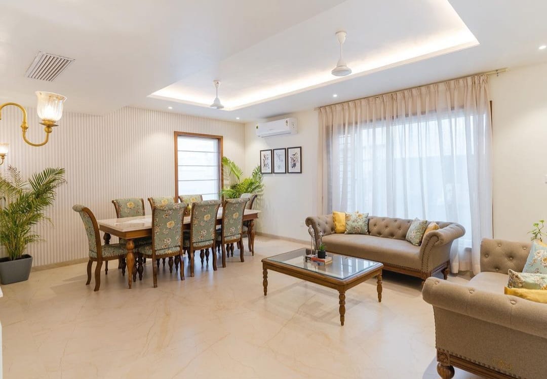 A spacious, elegantly designed dining room featuring an 8-seater marble top dining set with pastel chairs, accompanied by sophisticated living room furniture and decorative lighting.