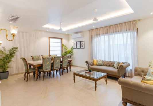 A spacious, elegantly designed dining room featuring an 8-seater marble top dining set with pastel chairs, accompanied by sophisticated living room furniture and decorative lighting.