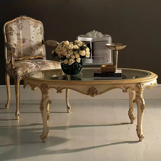 An ornate antique gold French oval coffee table with a glass top, featuring intricate carved details on the legs and sides, placed in a room with a floral chair and decorative items on top.