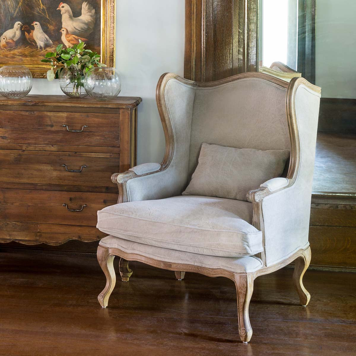 A beige upholstered high armchair with a distressed wooden frame, placed in a room with wooden flooring and a wooden side table.