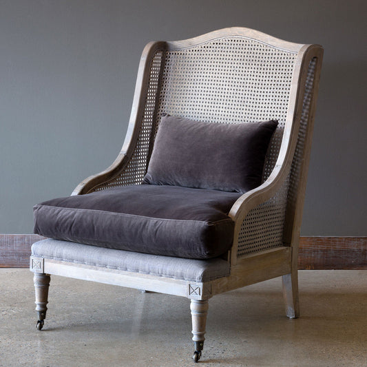A rustic grandeur cane armchair with light gray fabric seat and dark gray cushion, featuring intricate cane work and wooden legs with wheels.